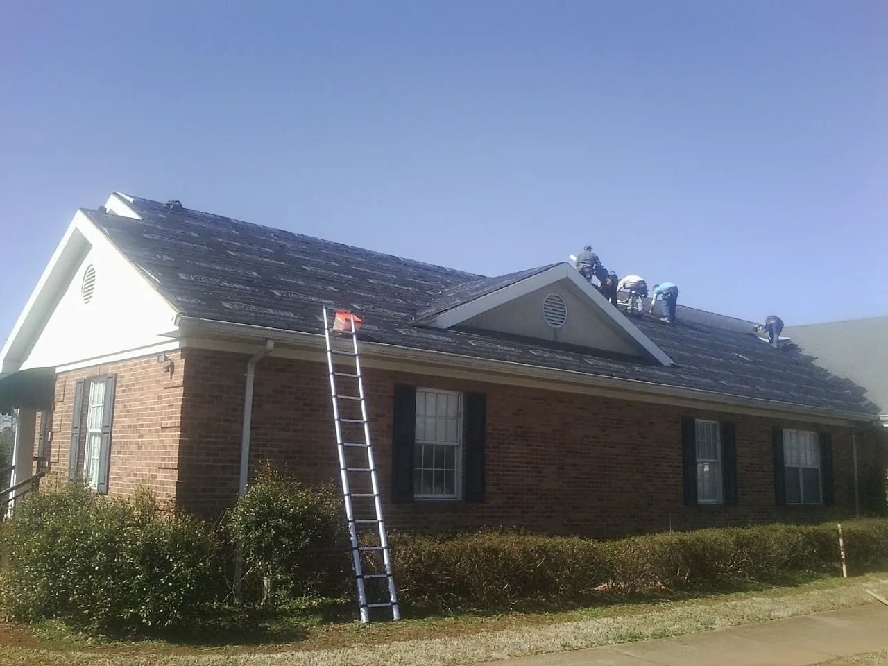 Crew tearing shingles off a brick ranch roof with a ladder set — Emergency Roof Repair work by our crew in Dutton